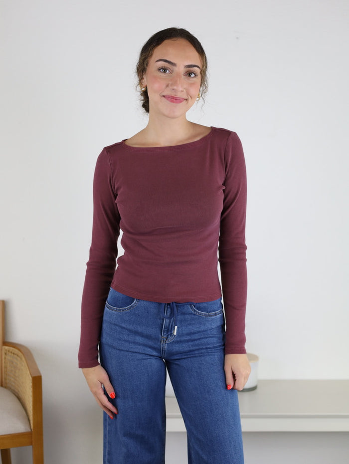 A woman with curly hair in a bun wears the Clove Fitted Rib Knit Boat Neck Top in maroon and blue jeans, standing and smiling in a simple room with a chair and small table in the background.