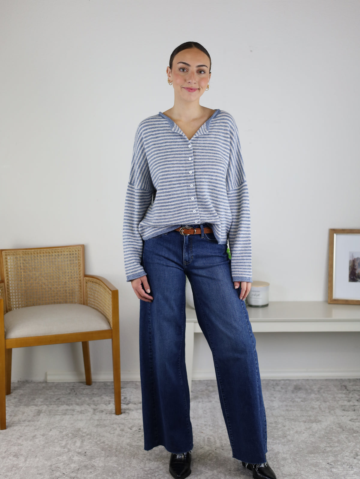 A woman stands indoors wearing the Maren Striped Cardigan—a relaxed fit gray and white striped sweater—paired with wide-leg blue jeans. She is next to a wooden chair and a white bench with decor, against a plain light wall.