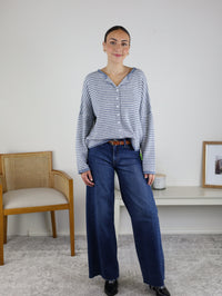 A woman stands indoors wearing the Maren Striped Cardigan—a relaxed fit gray and white striped sweater—paired with wide-leg blue jeans. She is next to a wooden chair and a white bench with decor, against a plain light wall.