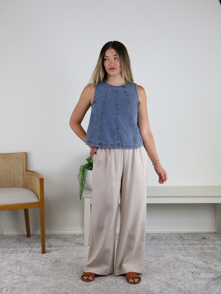 A woman stands indoors wearing the Rebecca Seam Detail Sleeveless Denim Top – Final Sale, styled as a versatile summer blouse and paired with wide-leg beige pants and brown sandals. She stands beside a wooden chair and a white bench topped with a small green plant, against a plain light wall backdrop.