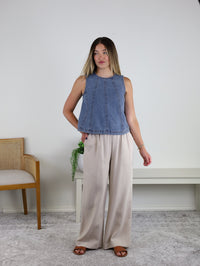 A woman stands indoors wearing the Rebecca Seam Detail Sleeveless Denim Top – Final Sale, styled as a versatile summer blouse and paired with wide-leg beige pants and brown sandals. She stands beside a wooden chair and a white bench topped with a small green plant, against a plain light wall backdrop.