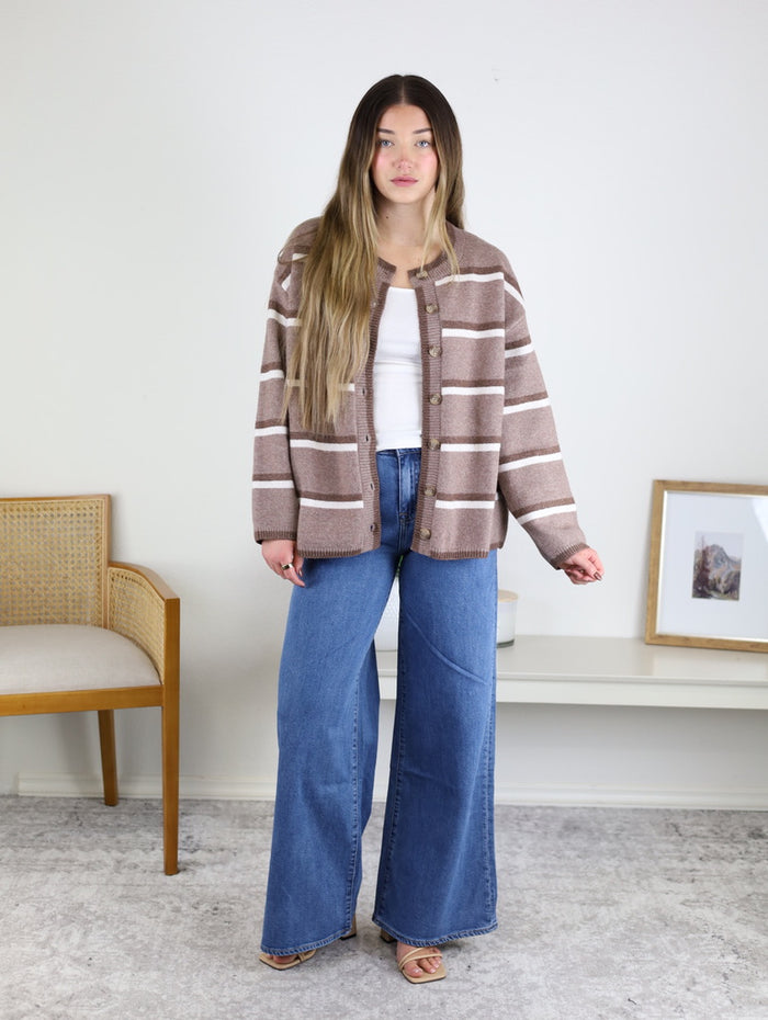 A person wears a brown and white Nora Stripe Button Down Knit Cardigan over a white tank top with blue wide-leg jeans.