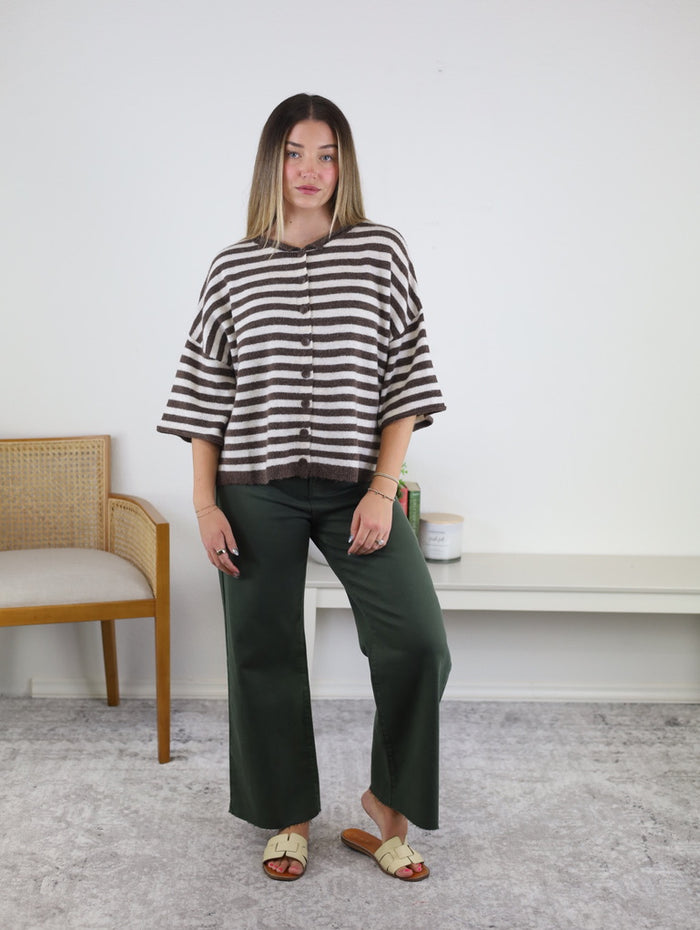 A woman stands indoors wearing a loose striped top, the Anita Scissor Cut Wide Leg Jean in a dark olive wash with a raw-hem denim finish, and beige sandals. She has straight, shoulder-length hair and poses in front of a white wall with minimal furniture.