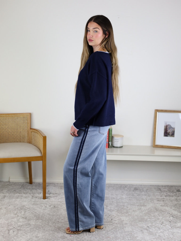 Woman with long hair wears the Bell Contrast Boat Neck Sweater and wide-leg jeans with a dark side stripe, standing sideways in a minimalist room with a woven chair, low shelf, framed picture, and candle.