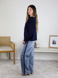 Woman with long hair wears the Bell Contrast Boat Neck Sweater and wide-leg jeans with a dark side stripe, standing sideways in a minimalist room with a woven chair, low shelf, framed picture, and candle.