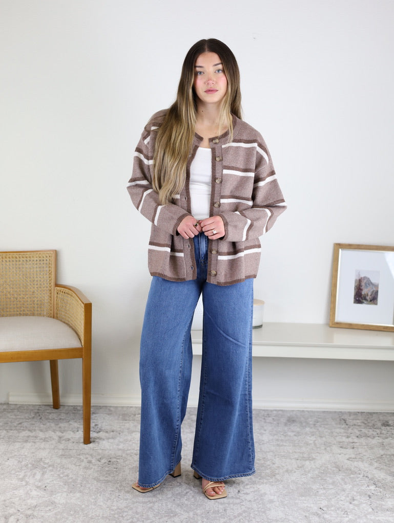 A person wears a brown Nora Stripe Button Down Knit Cardigan over a white top and wide-leg blue jeans.