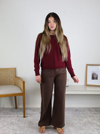 A woman wears a burgundy viscose blend Eli Front Seam Sweater with a vertical front seam and wide-leg brown trousers.