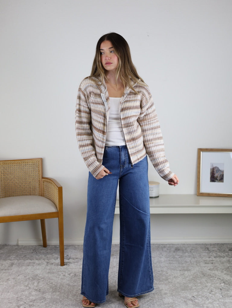 A person wears a brown and white striped Romy Hooded Cardigan Sweater over a white tank top and wide-leg blue jeans.