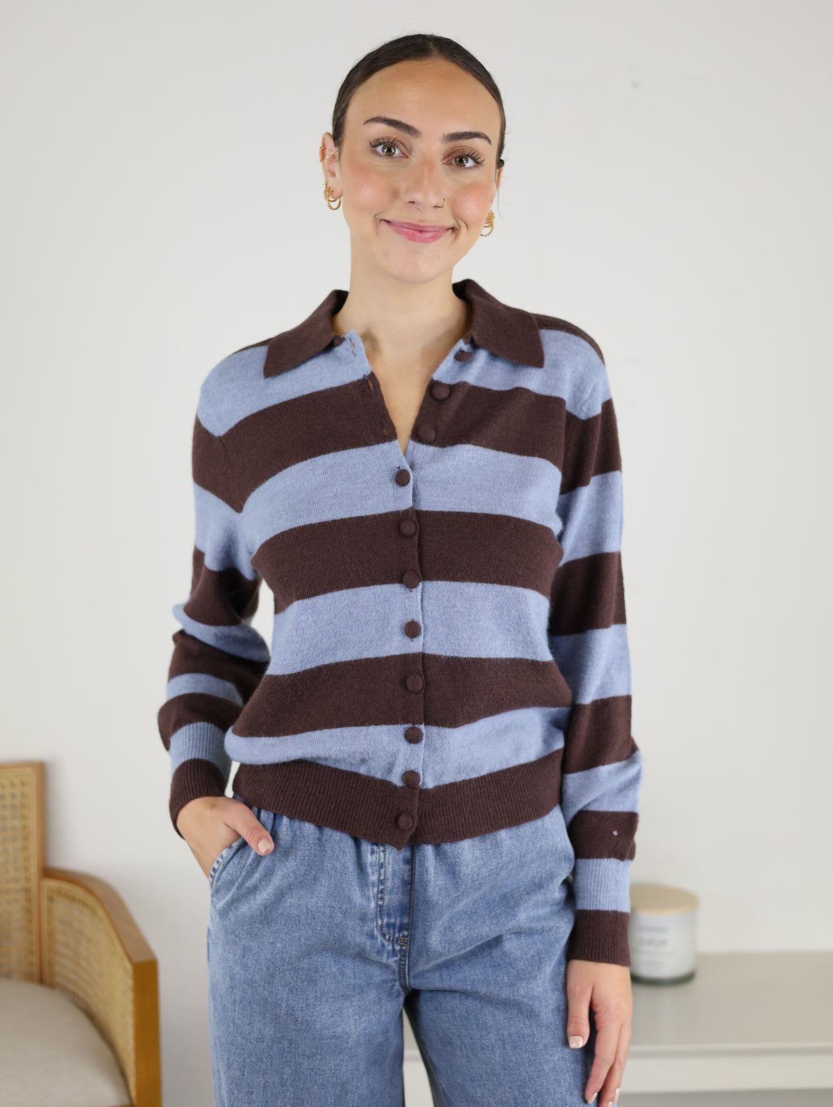 A woman stands indoors, smiling, in the Sloane Wide Stripe Collared Cardigan featuring cozy brown and light blue stripes, paired with blue jeans and her left hand in her pocket. Behind her is a light-colored wall, a chair, and a candle on a small table.