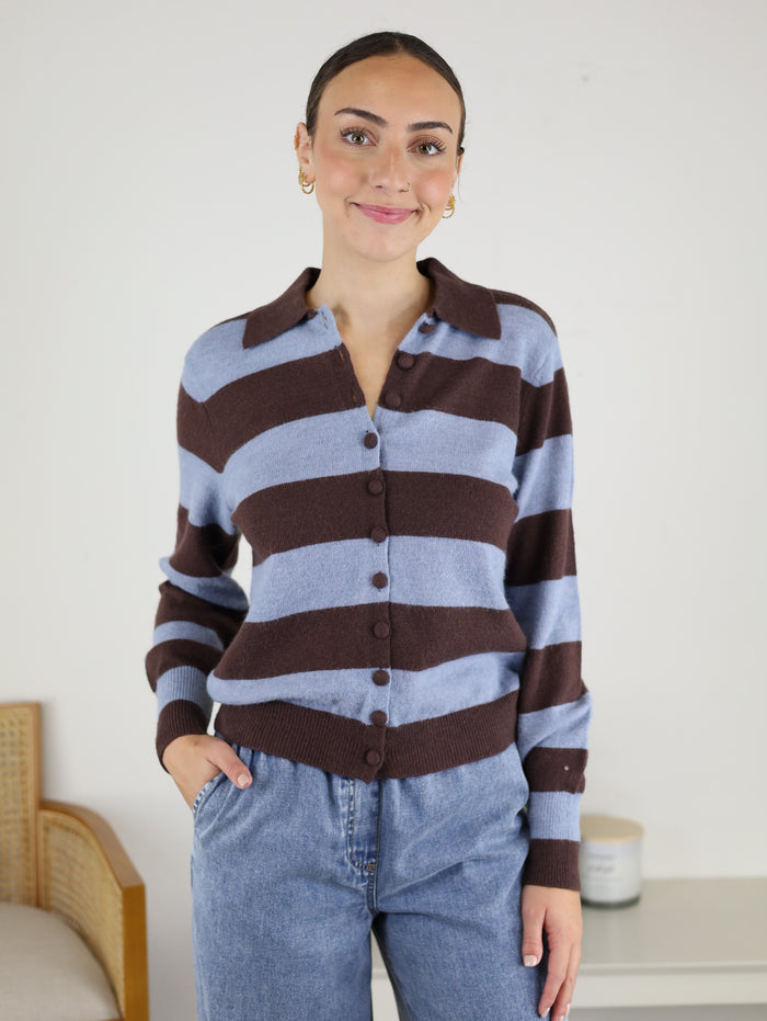 A woman stands indoors, smiling, in the Sloane Wide Stripe Collared Cardigan featuring cozy brown and light blue stripes, paired with blue jeans and her left hand in her pocket. Behind her is a light-colored wall, a chair, and a candle on a small table.