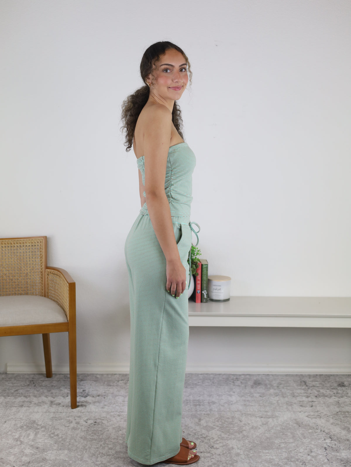 A woman in a minimal room with a chair, shelf, books, and a plant stands sideways, smiling in the Kia Striped Tube Top and Pants Set—a light green strapless jumpsuit that brings fresh style to her look.