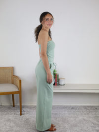 A woman in a minimal room with a chair, shelf, books, and a plant stands sideways, smiling in the Kia Striped Tube Top and Pants Set—a light green strapless jumpsuit that brings fresh style to her look.
