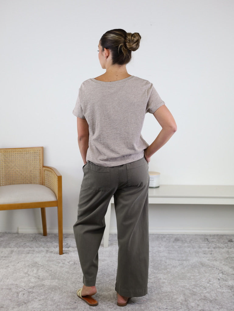 A woman stands indoors with her back to the camera, wearing the Nomad V Neck Tee in beige, crafted from soft knit fabric. She pairs it with gray wide-leg pants and flats, her hair styled in a bun. A wooden chair and white shelf in the background complement the relaxed fit of her effortlessly chic look.