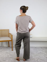 A woman stands indoors with her back to the camera, wearing the Nomad V Neck Tee in beige, crafted from soft knit fabric. She pairs it with gray wide-leg pants and flats, her hair styled in a bun. A wooden chair and white shelf in the background complement the relaxed fit of her effortlessly chic look.