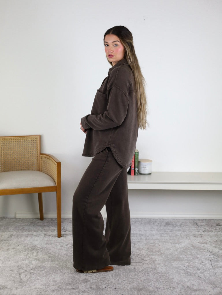Woman with long hair stands sideways in a minimalist room—with a cane chair, small shelf, books, and candles in the background—wearing the Hunter Knit Pant in Dark Chocolate.