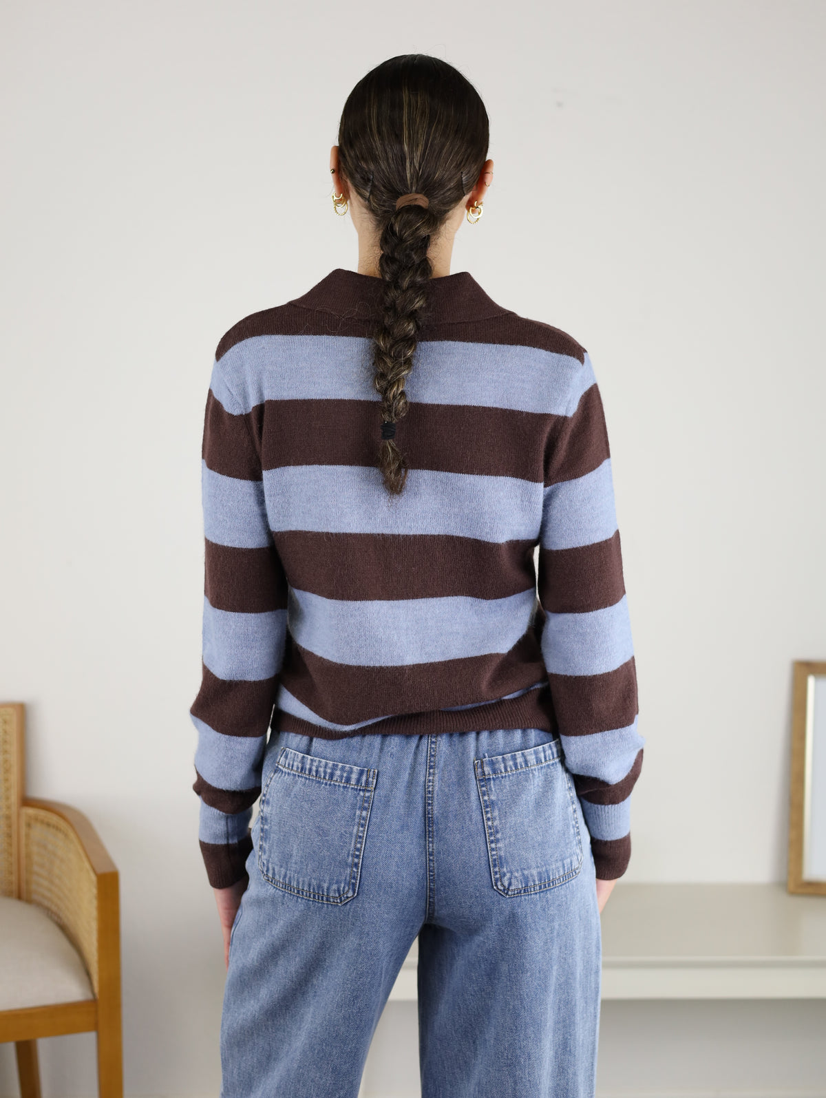 Wearing the Sloane Wide Stripe Collared Cardigan in brown and blue stripes with blue jeans, a person with a single braid stands with their back to the camera in a simple room furnished with a chair and table.