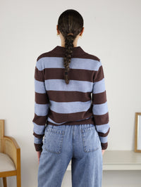 Wearing the Sloane Wide Stripe Collared Cardigan in brown and blue stripes with blue jeans, a person with a single braid stands with their back to the camera in a simple room furnished with a chair and table.
