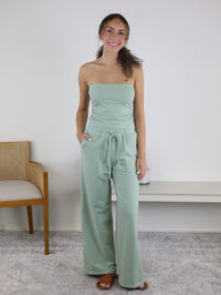 A woman with curly hair smiles while wearing the Kia Striped Tube Top and Pants Set, which features a light green striped strapless top paired with women’s wide-leg pants. She stands indoors on a gray rug, with a chair and a white bench in the background.