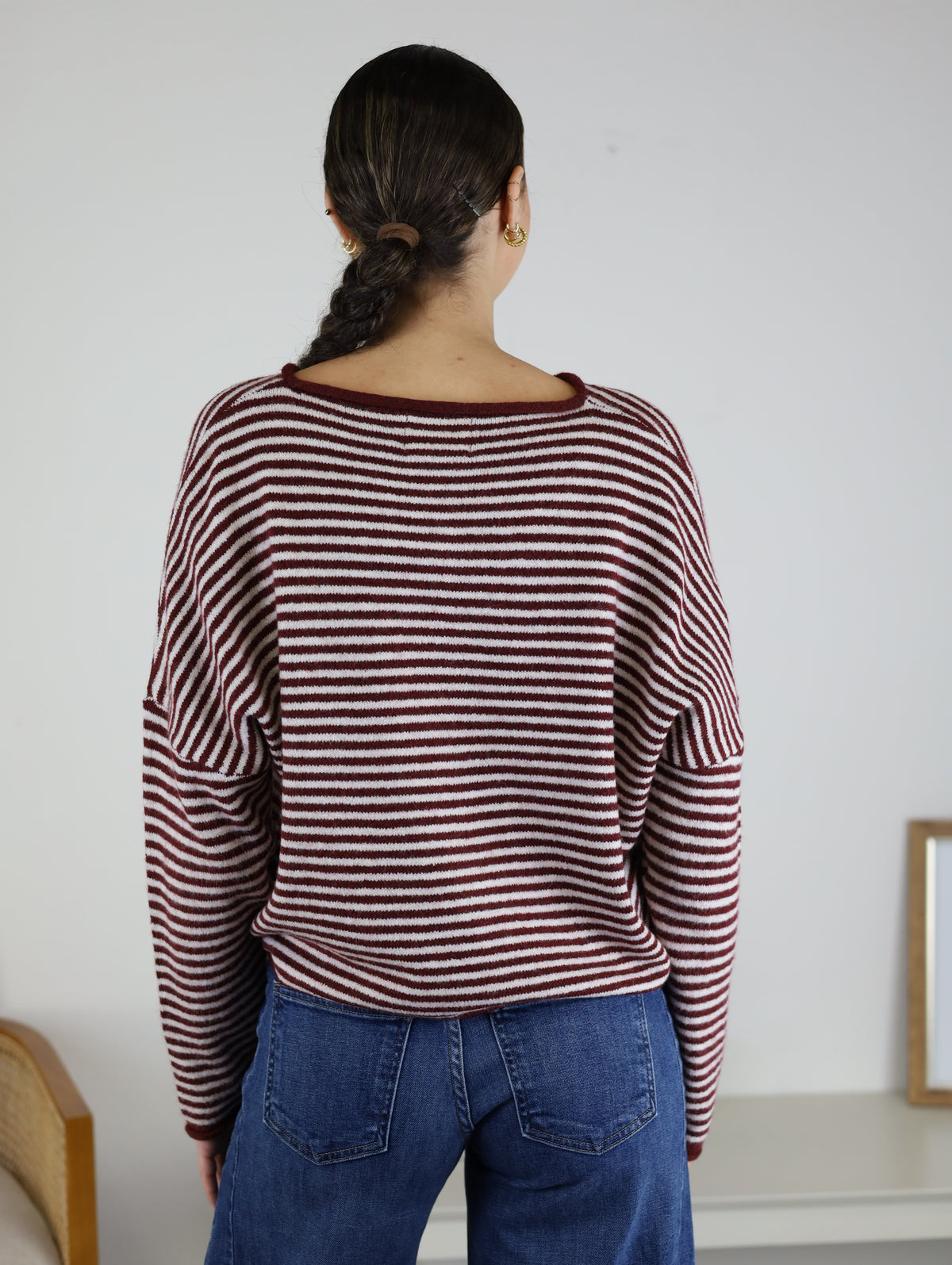 A person with long dark hair in a braid, wearing the Maren Striped Cardigan—a relaxed fit red and white striped cardigan—paired with blue jeans, stands facing away from the camera in a minimalist room.