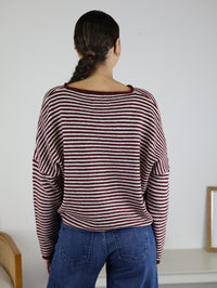 A person with long dark hair in a braid, wearing the Maren Striped Cardigan—a relaxed fit red and white striped cardigan—paired with blue jeans, stands facing away from the camera in a minimalist room.