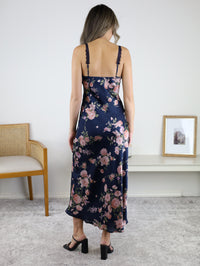 A woman seen from the back wearing a dark blue Kameron Floral Lace Trim Maxi Dress with lace-trimmed spaghetti straps and black block-heeled sandals.