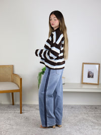 A person wears a Remy Multicolor Rolled Neck Knit Cardigan featuring a brown, white, and blue stripe pattern and a rolled neck, paired with light-wash jeans featuring a dark side stripe.