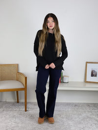 A woman with long, wavy hair wears a black ribbed knit Finley Sweater, navy pants, and brown shoes, standing indoors by a white wall near a wooden chair and minimalist shelf with decor.