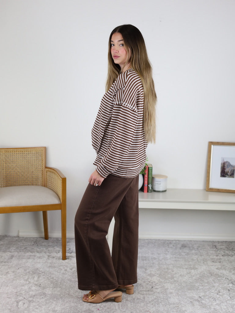 A woman with long hair models an oversized Lennon Stripe Knit Top with wide-leg brown pants and heeled sandals indoors, next to a chair, shelf, photo frame, and candles in the background.