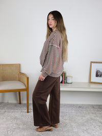A woman with long hair models an oversized Lennon Stripe Knit Top with wide-leg brown pants and heeled sandals indoors, next to a chair, shelf, photo frame, and candles in the background.