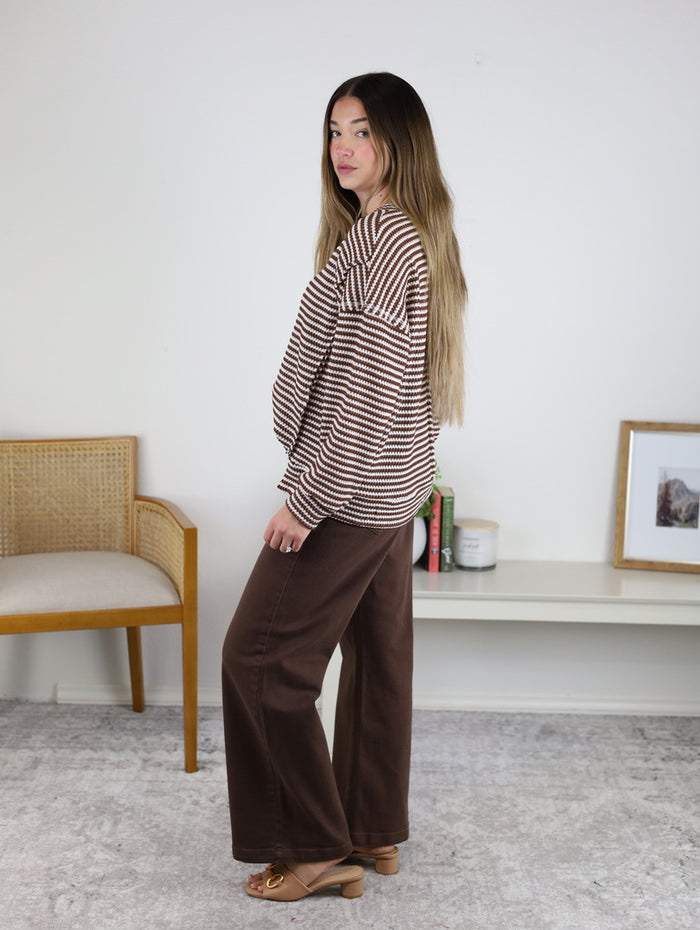 A woman with long hair models an oversized Lennon Stripe Knit Top with wide-leg brown pants and heeled sandals indoors, next to a chair, shelf, photo frame, and candles in the background.