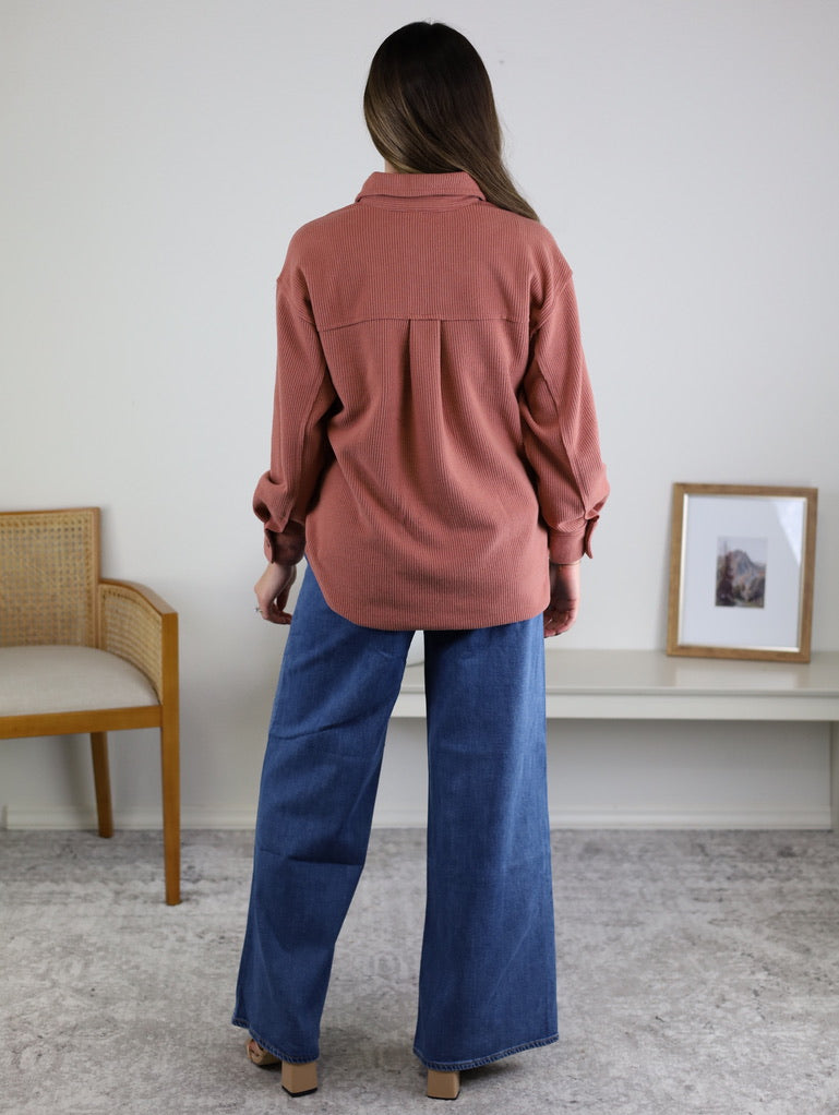 A woman stands indoors facing away, wearing the Larkin Rib Knit Button Up in rose and wide-leg blue jeans. A wooden chair, small bench, and framed picture are in the background.