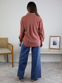 A woman stands indoors facing away, wearing the Larkin Rib Knit Button Up in rose and wide-leg blue jeans. A wooden chair, small bench, and framed picture are in the background.