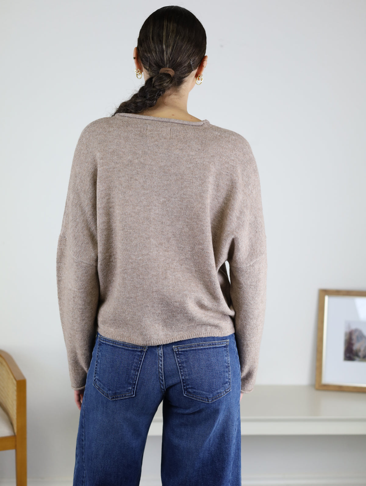 A person with dark hair in a braid, wearing the Beth Lightweight Cardigan in light brown and blue jeans, stands with their back to the camera in a minimal room featuring a chair and a framed photo on a white table.