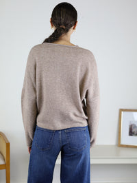 A person with dark hair in a braid, wearing the Beth Lightweight Cardigan in light brown and blue jeans, stands with their back to the camera in a minimal room featuring a chair and a framed photo on a white table.