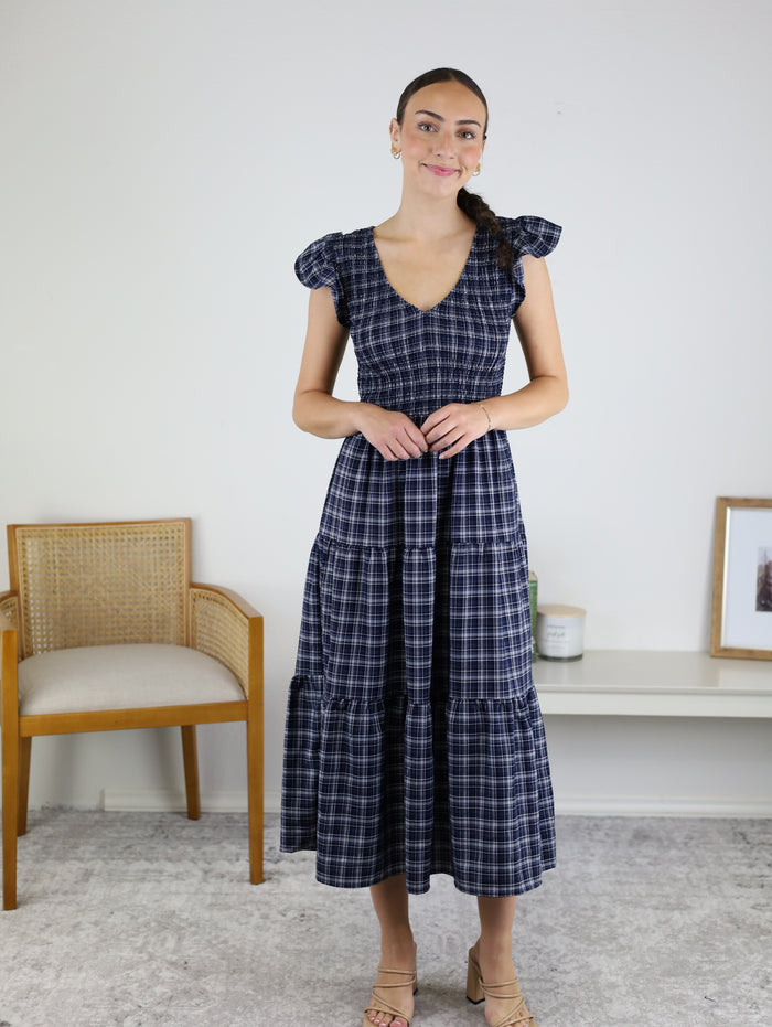 A woman wears the Brynn Plaid V-Neck Tiered Midi Dress as she stands indoors on a light rug beside a wooden chair and a white shelf adorned with decor. Smiling softly with her hair in a side braid, she embodies effortless fall style in this navy plaid tiered dress.