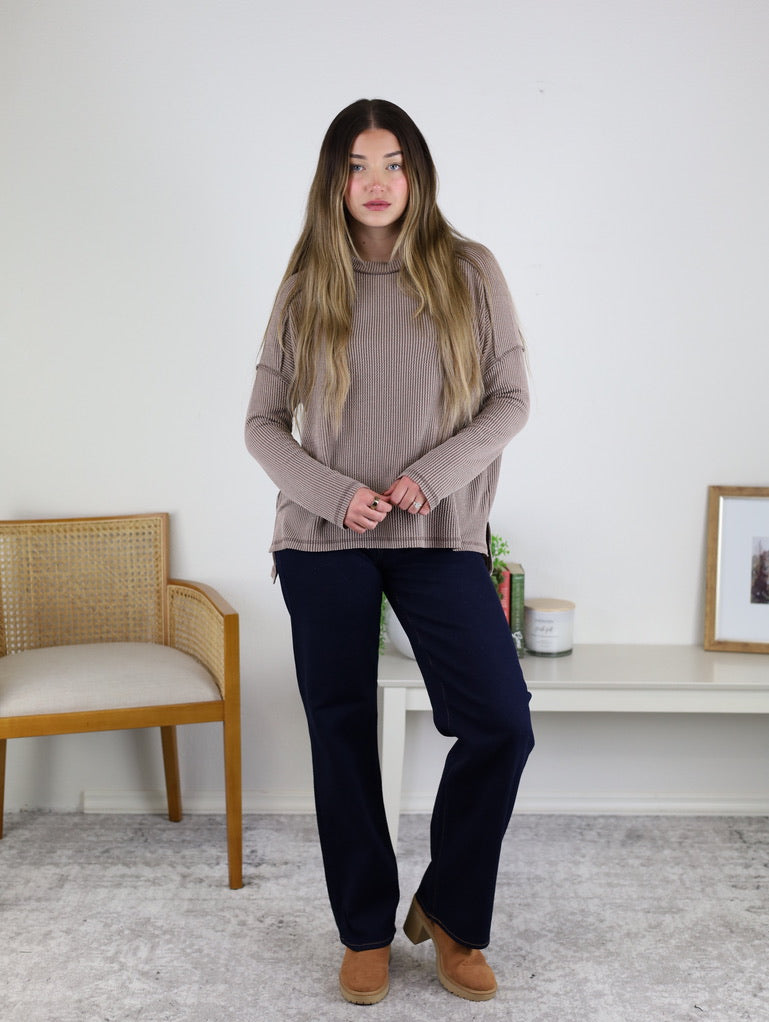 A woman with long brown hair stands indoors wearing the Finley Sweater, dark pants, and brown shoes beside a wooden chair and a white table holding books, a candle, and a framed picture.