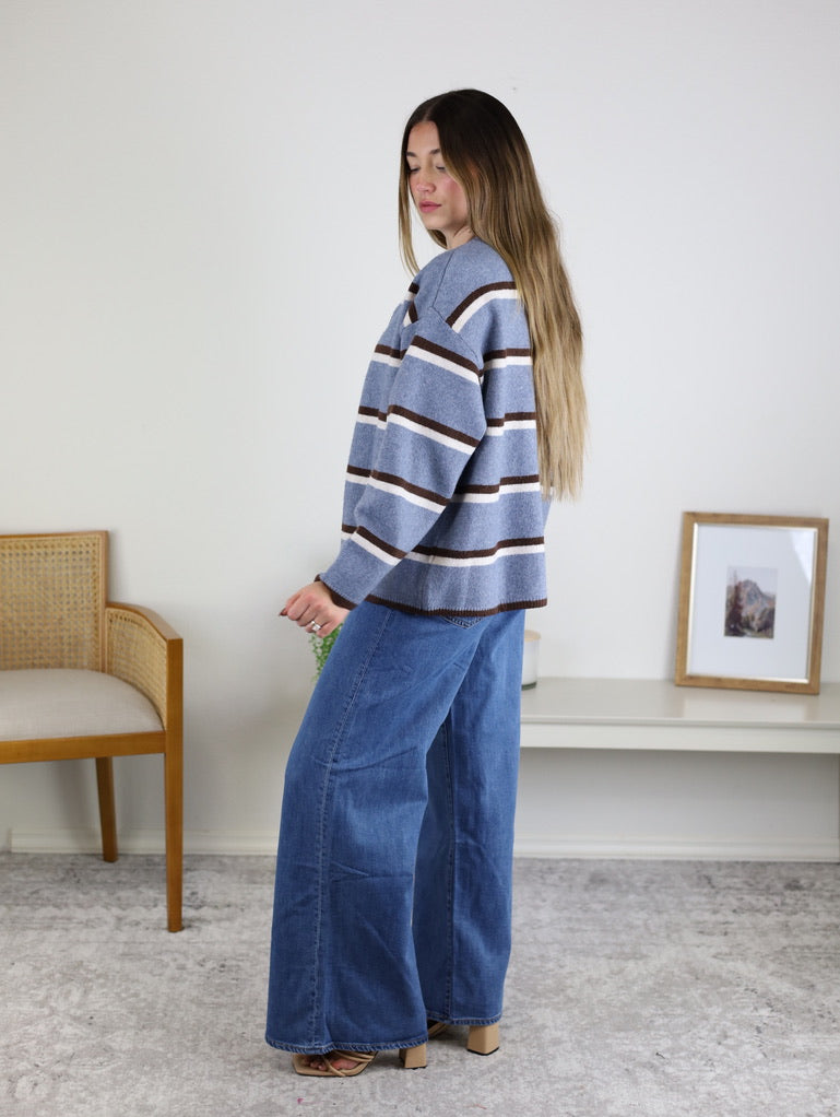A woman wears the Nora Stripe Button Down Knit Cardigan, a light blue knit sweater with brown and white horizontal stripes, paired with wide-leg blue jeans.