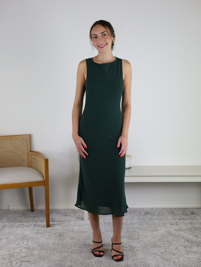 A woman in the Rudi Boat Neck Cowl Back Midi Dress and black sandals stands indoors, smiling. She is next to a wooden chair with a cushion and a white bench, against a light wall and gray carpet.