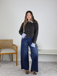 A woman stands indoors wearing the Mani Stripe Trim Button Down Cardigan over a blue shirt and wide-leg jeans with black sandals. A wooden chair and a bench with plants are in the background against a white wall.