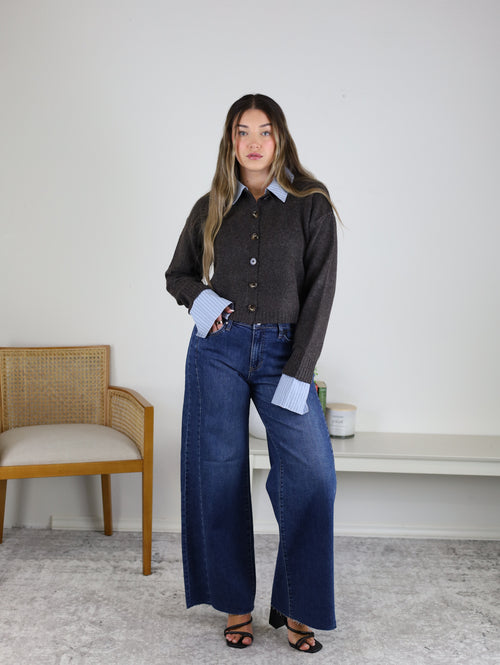 A woman stands indoors wearing the Mani Stripe Trim Button Down Cardigan over a blue shirt and wide-leg jeans with black sandals. A wooden chair and a bench with plants are in the background against a white wall.