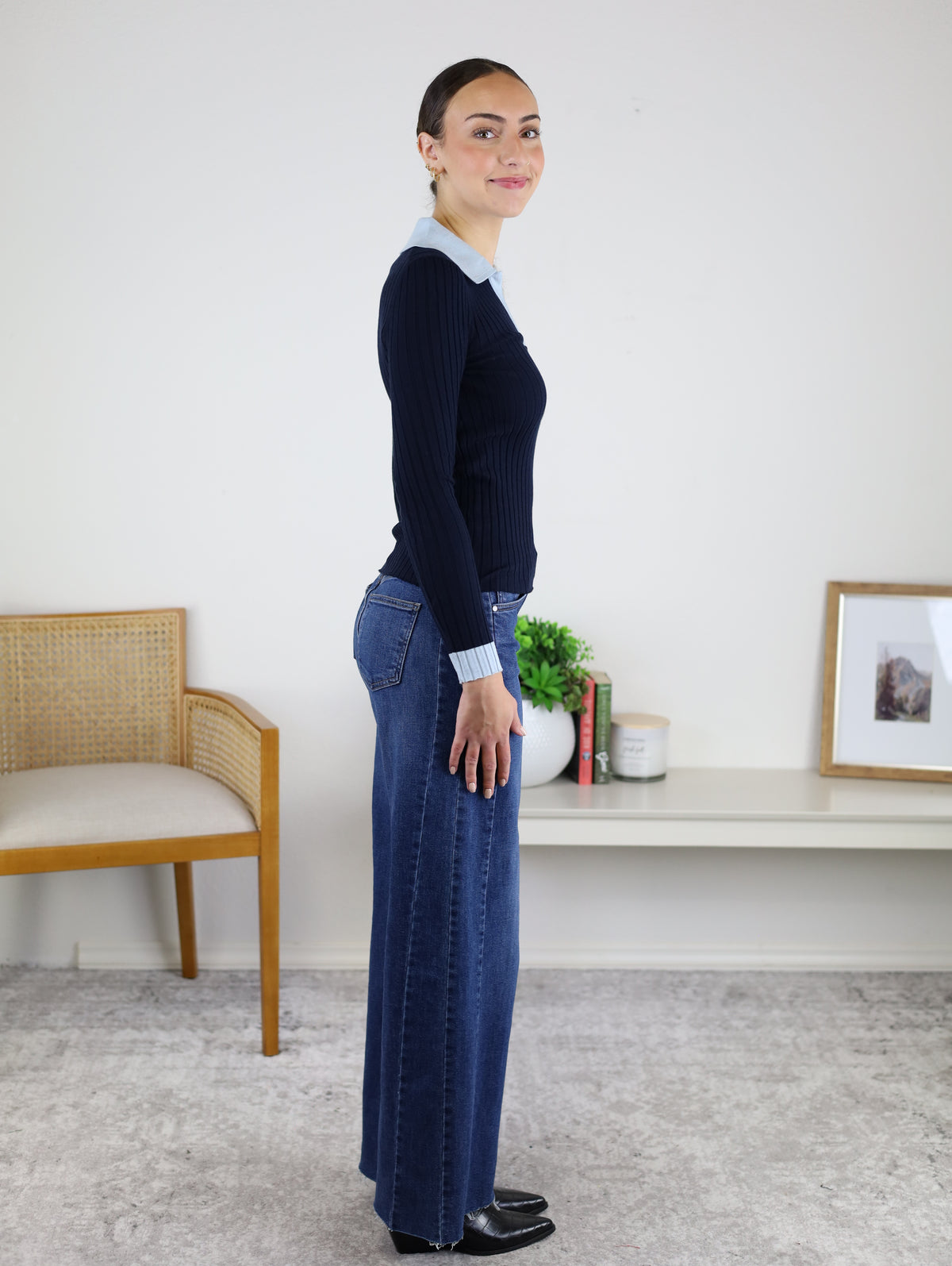 A woman stands in profile indoors, wearing the Deepsea Ribbed Collared Knit Top in black with blue jeans and black shoes. Behind her are a wooden chair, a small table with decor, and framed art on the wall.