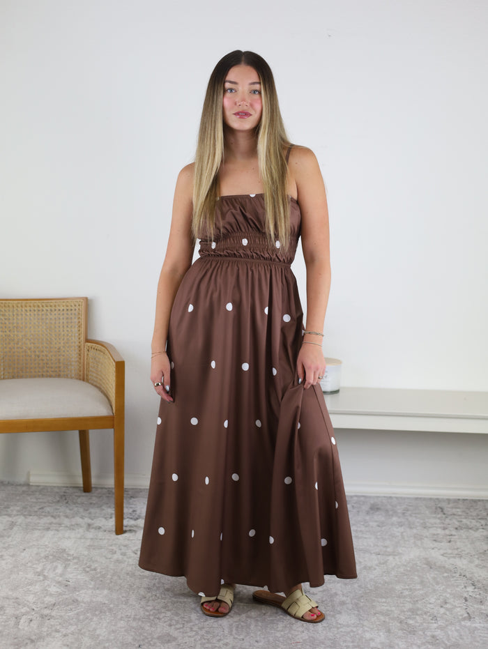Wearing the Reverie Polka Dot Maxi Dress, a woman stands indoors on a light gray carpet, her summer-ready look completed with beige sandals. The strapless brown maxi dress features white polka dots and is effortlessly complemented by the wooden chair and white bench in the background—perfect attire for a vacation getaway.