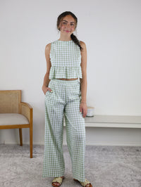 A woman stands indoors wearing the Myra Gingham Print Ruffled Set, which features a light green and white checkered sleeveless crop top and matching high-waisted wide-leg pants. She is smiling with one hand relaxed by her side, while a chair and a bench are visible in the background.
