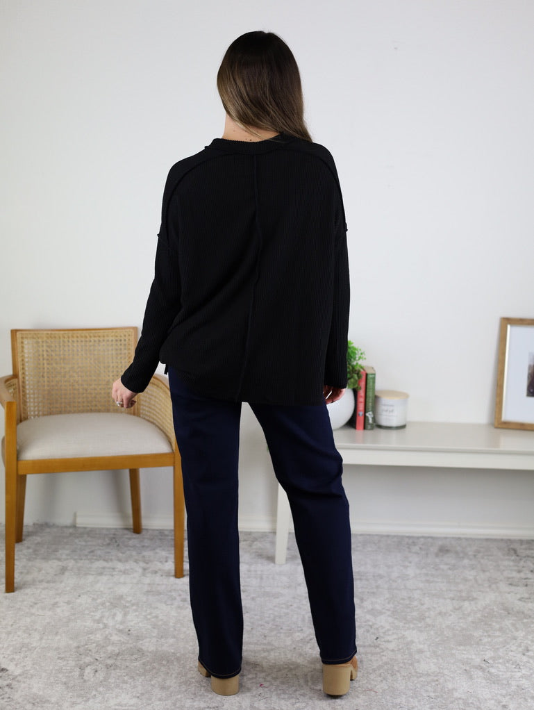 A woman with long brown hair stands indoors facing away, wearing the Finley Sweater—a ribbed black knit—paired with navy pants and tan shoes, near a wooden chair and decor on a white shelf.