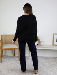 A woman with long brown hair stands indoors facing away, wearing the Finley Sweater—a ribbed black knit—paired with navy pants and tan shoes, near a wooden chair and decor on a white shelf.