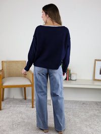 A woman stands indoors facing away, wearing the Bell Contrast Boat Neck Sweater in navy with light blue jeans. She’s in a neutral room with a wooden chair, white shelf, books, and framed art in the background.