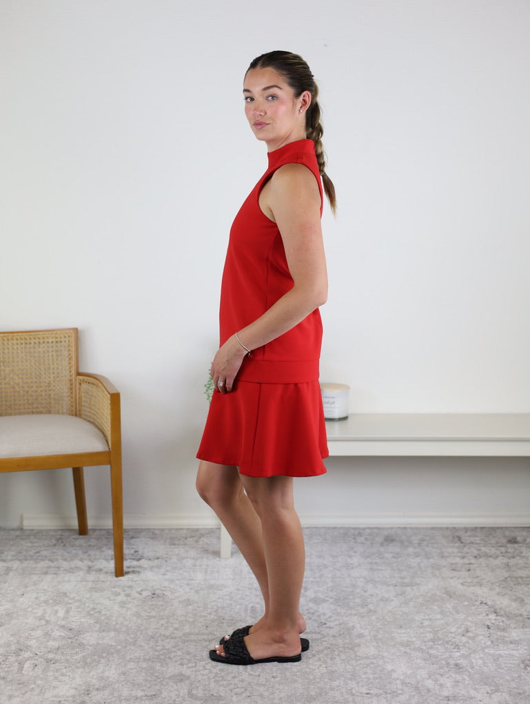 A young woman with a braided hairstyle stands in profile, wearing the Cooper Sleeveless Mock Neck Shift Dress in vibrant red, paired with black sandals. She is indoors beside a wooden chair and a white bench against a plain white wall.