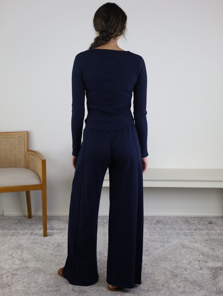 A woman with long brown hair in a braid stands facing away in a minimal room with a beige chair and a white bench, wearing the Statler Lounge Set—a navy blue henley top paired with wide-leg pants.