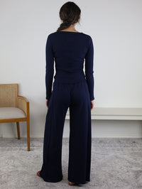 A woman with long brown hair in a braid stands facing away in a minimal room with a beige chair and a white bench, wearing the Statler Lounge Set—a navy blue henley top paired with wide-leg pants.