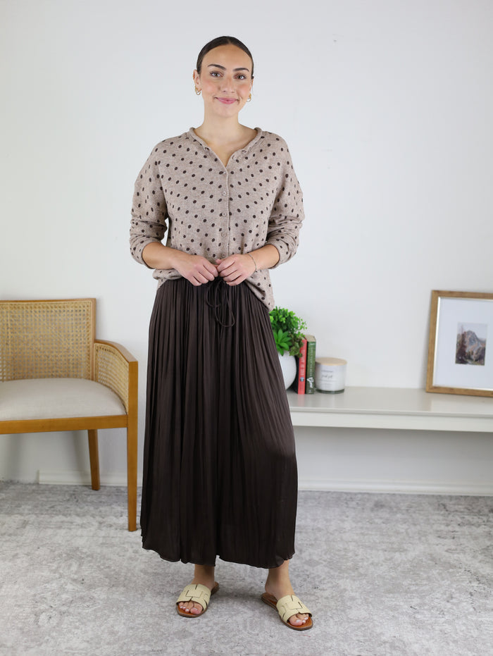A woman stands indoors wearing a beige polka-dot blouse, sandals, and the Maci Satin Crinkle Pleated Maxi Skirt in a dark hue. Smiling as she holds her skirt with one hand, she poses before a backdrop of a chair, shelf, books, and framed photo—making this elegant skirt an ideal addition to any fall wardrobe.
